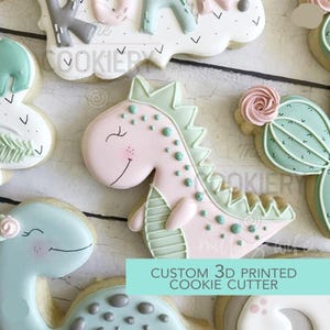 May include: Assortment of decorated sugar cookies in the shape of dinosaurs, a cactus, and a cloud. The cookies are decorated with pastel pink, green, and blue icing. The text "CUSTOM 3D PRINTED COOKIE CUTTER" is visible.