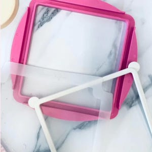 May include: A pink square embroidery hoop with a clear plastic insert and a white frame. The hoop is made of plastic and has a rounded shape on the corners. The white frame is designed to hold the fabric in place.