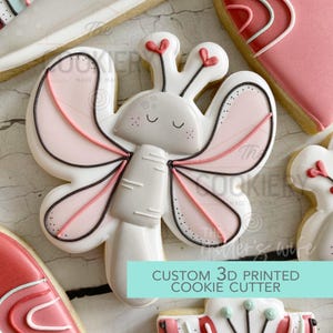 May include: A butterfly-shaped cookie cutter with a white base, pink and black details, and a smiling face. The wings have pink and black accents. The text "CUSTOM 3D PRINTED COOKIE CUTTER" is visible.