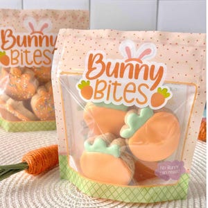 May include: Two clear plastic bags of Easter cookies. The bags are labeled "Bunny Bites" in orange and white. One bag contains carrot-shaped cookies with green frosting. The other bag contains bunny-shaped cookies with sprinkles.
