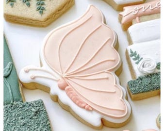 May include: A collection of decorated sugar cookies. A large butterfly-shaped cookie with pink icing and white trim is the focal point. Other cookies include floral and geometric designs, with green and white icing.