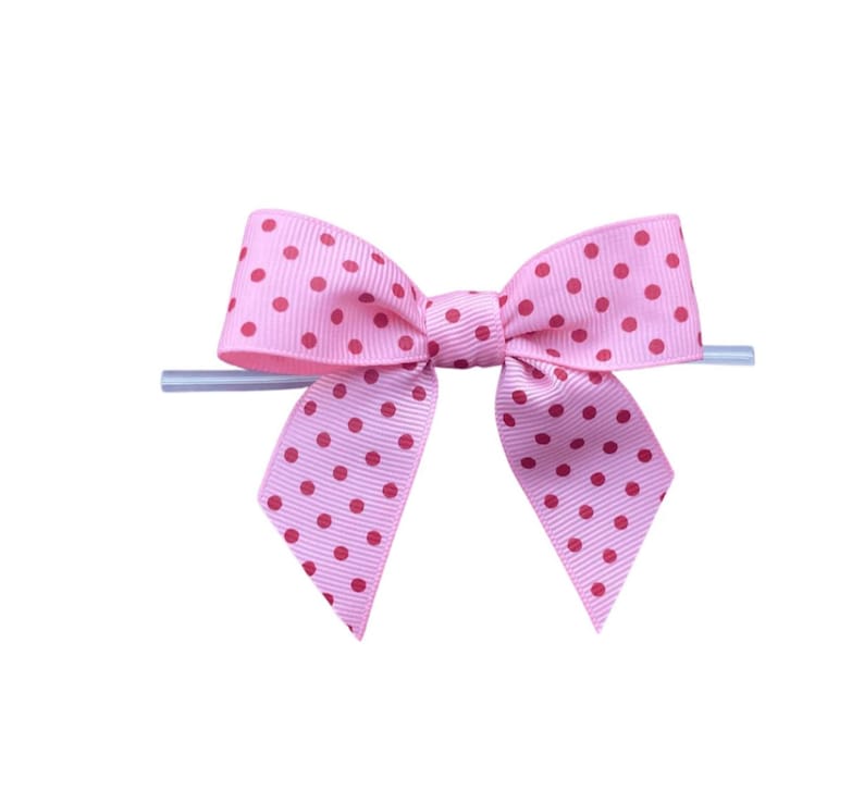 Op de afbeelding: Een roze strik met rode stippen. De strik is gemaakt van lint en heeft een klassieke vorm. Het is bevestigd aan een doorzichtige plastic stok, waarschijnlijk voor weergave of bevestiging.