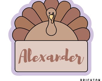 May include: A turkey-shaped cookie cutter with a light brown body and a fan of brown feathers. The name "Alexander" is written in a cursive font on the bottom. The cutter has a purple outline and the words "BRIGHTON Cutters" are at the bottom.