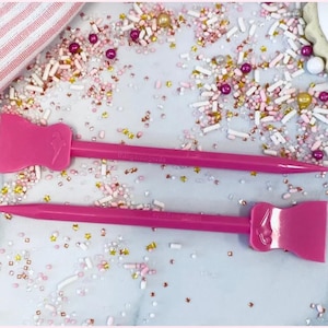 May include: Two bright pink icing spatulas with a pointed end and a flat, spatula-like end. The text "thingamagenta" is printed on the handle of each spatula. The spatulas are surrounded by colorful sprinkles.