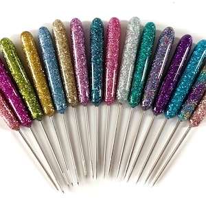 May include: A collection of decorative pins with glitter-covered handles in various colors, including pink, green, gold, blue, and purple. The pins have long, thin, silver metal shafts and are arranged in a fan shape against a white background.