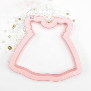 May include: A light pink cookie cutter in the shape of a dress. The cutter has a detailed design with a bodice and skirt. The letters "KZ" are visible on the top of the cutter. Sprinkles and decorative beads surround the cutter.