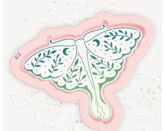 May include: A pink and white moth-shaped mold with a detailed design. The moth features a white body and wings with green and blue accents, including crescent moons and leaf patterns. The letters "KZ" are visible on the side.