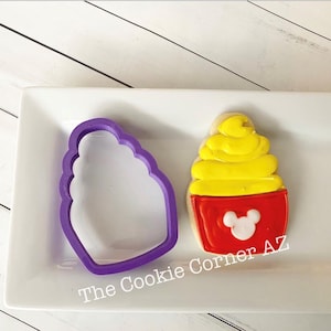 May include: A purple plastic cookie cutter and a decorated cookie shaped like a cupcake. The cookie has a red base with a white Mickey Mouse head and yellow frosting. The text "The Cookie Corner AZ" is visible.