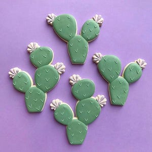 May include: Cactus-shaped cookies with green frosting and white accents, arranged on a purple surface. Each cookie features a detailed design, resembling a prickly pear cactus. The cookies are a light green color and have a delicate, handcrafted appearance.