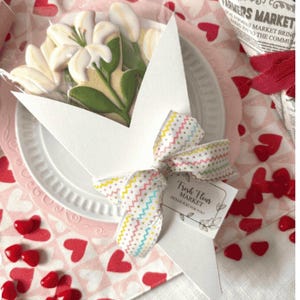 May include: A white plate holds flower-shaped cookies wrapped in clear cellophane, tied with a patterned ribbon and a tag that reads "Fresh Flour Market". The cookies are on a pink heart-patterned surface.