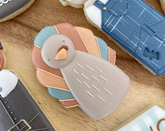 May include: A decorated sugar cookie in the shape of a turkey with a tan body, a light brown beak, and a colorful fan of feathers in shades of blue, orange, and brown. Other cookies in the image include a scarecrow and overalls.