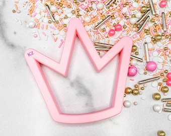 May include: A pink crown-shaped cookie cutter with the initials "KZ" on the side. The cutter is surrounded by a colorful assortment of sprinkles, including pink, white, gold, and silver, on a white surface.
