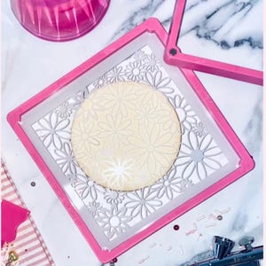 May include: A pink cookie stencil with a daisy pattern is centered over a round cookie. The stencil is held in place by a pink frame. Sprinkles and an airbrush are visible in the background.