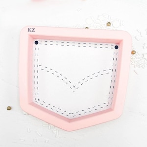 May include: A light pink cookie cutter in the shape of a pocket. The pocket design features a white interior with black dashed lines outlining the pocket shape and stitching details. The letters "KZ" are visible in the upper left corner.