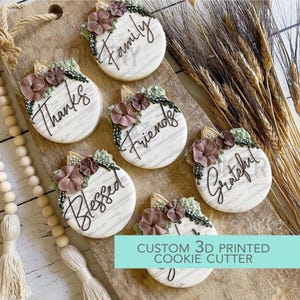 May include: Six round cookies with the words "Family", "Thanks", "Friends", "Blessed", and "Grateful" written in black script. Each cookie is decorated with brown flowers and green leaves. The text "CUSTOM 3D PRINTED COOKIE CUTTER" is at the bottom.