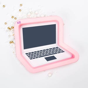 May include: A pink laptop-shaped object with a white keyboard and a dark gray screen. The object is surrounded by a pink border and sits on a white surface with gold and white decorative sprinkles.