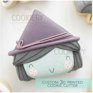 May include: A custom 3D printed cookie cutter in the shape of a witch's face. The witch has a lavender hat, gray hair, and a light blue face with pink cheeks. The text "The Cookery" is visible.