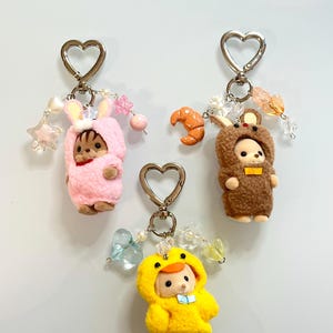 May include: Three keychains featuring small flocked animal figures in fuzzy costumes. One wears a pink rabbit suit, one a brown bear suit, and one a yellow duck suit. Each keychain has a silver heart-shaped clasp and assorted decorative beads.