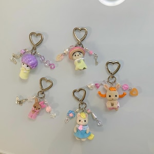 May include: Five keychains featuring small flocked animal figures, including a purple-haired character, a rabbit in a hat, a brown rabbit, a cat in a blue dress, and a rabbit with orange ears. Each keychain has a heart-shaped ring and beaded charms.