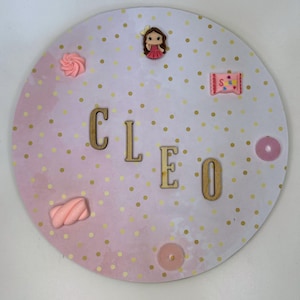 Può includere: Un cartello rotondo in legno rosa e bianco con punti di glitter dorati. Il cartello dice "CLEO" in lettere di legno e ha varie decorazioni rosa, tra cui una piccola bambola, una barretta di cioccolato e un marshmallow.