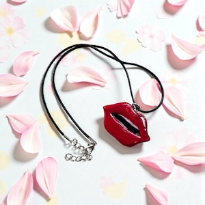 May include: A red lips pendant necklace with a black cord and silver-tone clasp. The pendant is glossy and detailed, set against a backdrop of pink flower petals and blossoms.