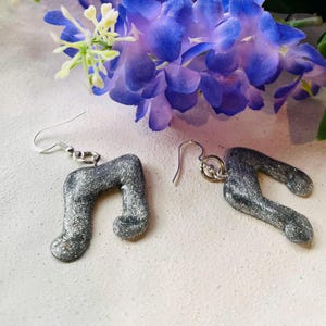 May include: A pair of silver glitter musical note earrings. The earrings are shaped like quaver notes and have silver-coloured ear wires. The background includes purple and yellow flowers.