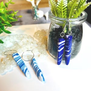 Può includere: Orecchini con design marmorizzato blu e bianco. Gli orecchini rettangolari presentano un motivo a spirale. Un paio è appeso a un barattolo di vetro, l'altro paio è su una superficie bianca. Ganci color argento.