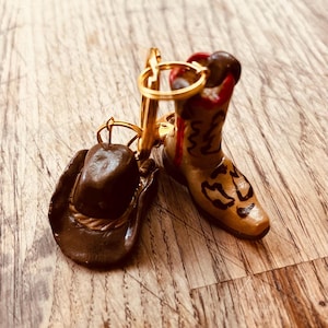 Puede incluir: Un llavero en miniatura con una bota y un sombrero de vaquero. La bota es de color beige con detalles en negro y marrón, y el sombrero es marrón. El llavero tiene un anillo y un cierre dorados. Los artículos están sobre una superficie de madera.