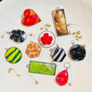 May include: A collection of ten assorted metal-framed pendants in various shapes including heart, rectangle, circle, hexagon, and teardrop. The designs feature diverse patterns and colors such as red, orange, gold, black, white, green, and yellow.