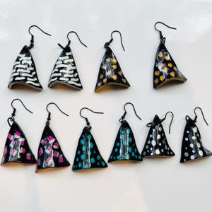 May include: A selection of handmade triangular earrings with black hooks. Each earring has a unique design, including white, pink, yellow, and blue dot and stripe patterns on a black base. The earrings are displayed on a white surface.