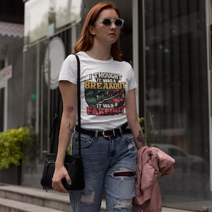 Puede incluir: Camiseta blanca con el texto "I thought it was a breakout, it was a fakeout" en negro, amarillo, rojo y verde. La camiseta se combina con vaqueros azules y un cinturón negro. Se ve un bolso negro al hombro.