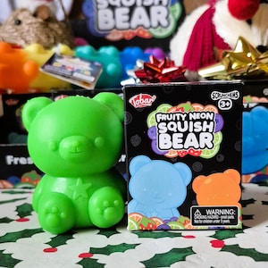 May include: A bright green squishy bear toy with a star on its chest sits next to a box. The box features the text "FRUITY NEON SQUISH BEAR" and illustrations of fruit and a blue bear. The toy is recommended for ages 3+.