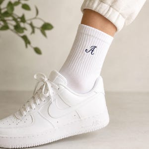 May include: White ribbed socks with a navy blue embroidered letter "A" are worn with white sneakers. The sneakers have white laces and a white sole. The socks are pulled up to the ankle.