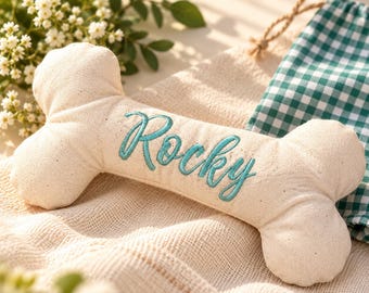 Puede incluir: Un juguete de tela para perro en forma de hueso de color crema, con el nombre Rocky bordado en letra cursiva de color verde azulado en el centro. El juguete está hecho de un material textil texturizado de tonos naturales con costuras visibles.