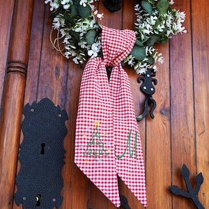 May include: Red and white gingham fabric with a green embroidered Christmas tree and the letter "M" on it. The fabric is hanging on a wooden door.