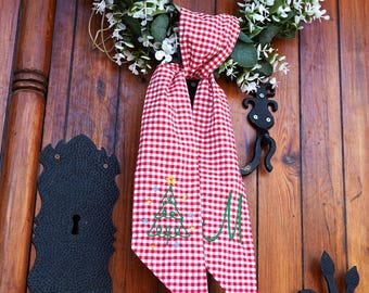 May include: Red and white gingham fabric with a green embroidered Christmas tree and the letter "M" on it. The fabric is hanging on a wooden door.