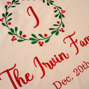 May include: Cream-colored fabric with a green wreath design and red berries. A red letter "I" is inside the wreath. The words "The Irwin Family" and "Dec. 20th" are embroidered in red script, suggesting a personalized holiday item.