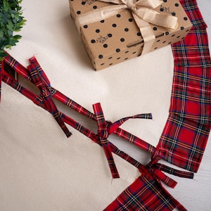 May include: A gift box with a gold ribbon bow sits on a cream-colored surface, next to a red and green plaid fabric. The box is brown with black polka dots and a logo. Red plaid ribbons are tied in a crisscross pattern.