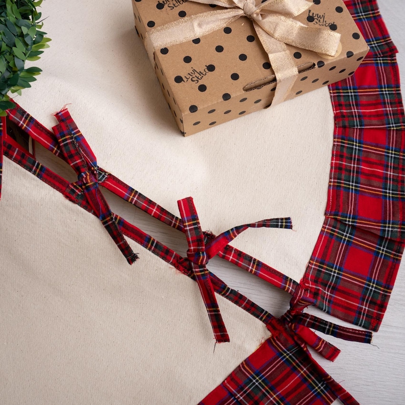 May include: A gift box with a gold ribbon bow sits on a cream-colored surface, next to a red and green plaid fabric. The box is brown with black polka dots and a logo. Red plaid ribbons are tied in a crisscross pattern.