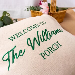 May include: Cream-colored pillow with green embroidered text that reads "WELCOME TO The Williams PORCH." A woven basket with flowers and greenery is in the background. The pillow is likely for outdoor decor.