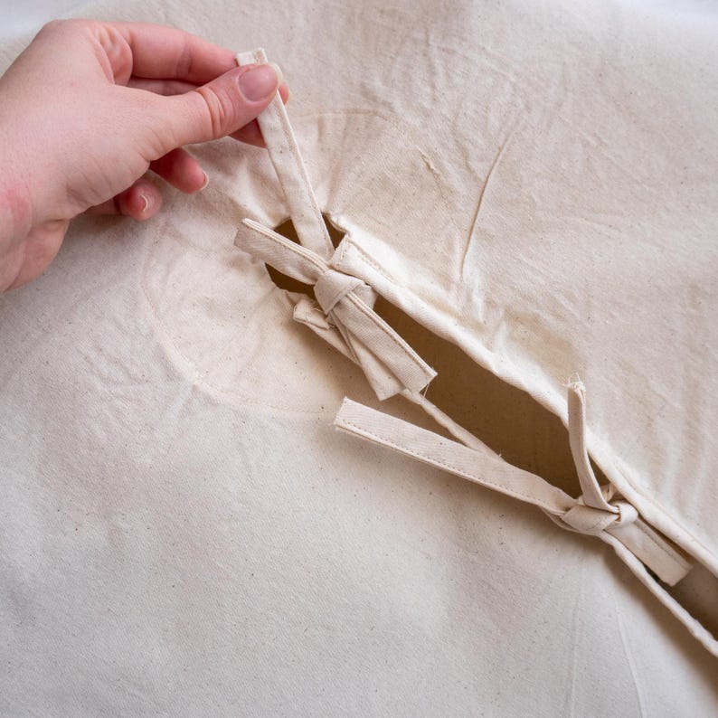 May include: Close-up of a cream-colored fabric with a slit and attached ties. The ties are knotted and appear to be made of the same fabric. A hand is holding one of the ties, suggesting a fastening mechanism or closure.