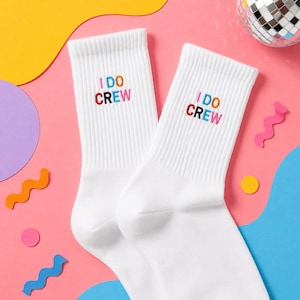 Könnte beinhalten: Ein Paar weiße Crew-Socken mit dem gestickten Text I DO CREW in mehrfarbigen Buchstaben. Die Socken haben eine gerippte Struktur am oberen Rand und bestehen aus weichem Stoff. Der Text ist in rosa, orange, blau, schwarz und rot gestickt.