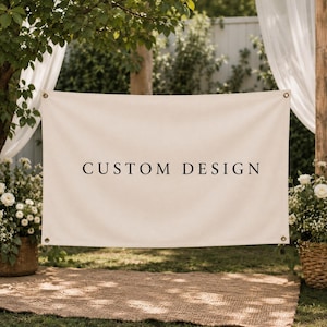 May include: A rectangular off-white fabric banner featuring the black text CUSTOM DESIGN centered on the front. The banner includes metal grommets in each of the four corners for hanging, providing a clean and neutral display surface.