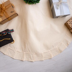 May include: A cream-colored Christmas tree skirt with a pleated edge. Several wrapped gifts in brown, silver, and black are scattered around the skirt. A small green wreath is at the top.