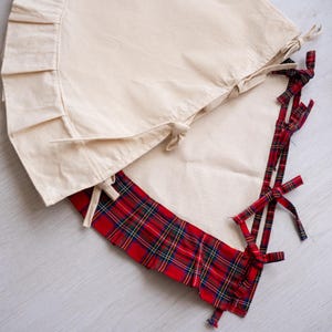 May include: Two-piece tree skirt. The top section is a solid cream color with a ruffled edge. The bottom section is red plaid with green and yellow accents. The skirt has cream-colored ties.