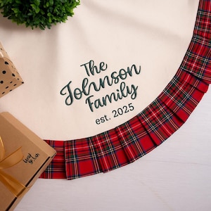 May include: Cream-colored Christmas tree skirt with a red and green plaid border. The skirt is embroidered with "The Johnson Family est. 2025" in green script. A gift box and greenery are visible in the background.