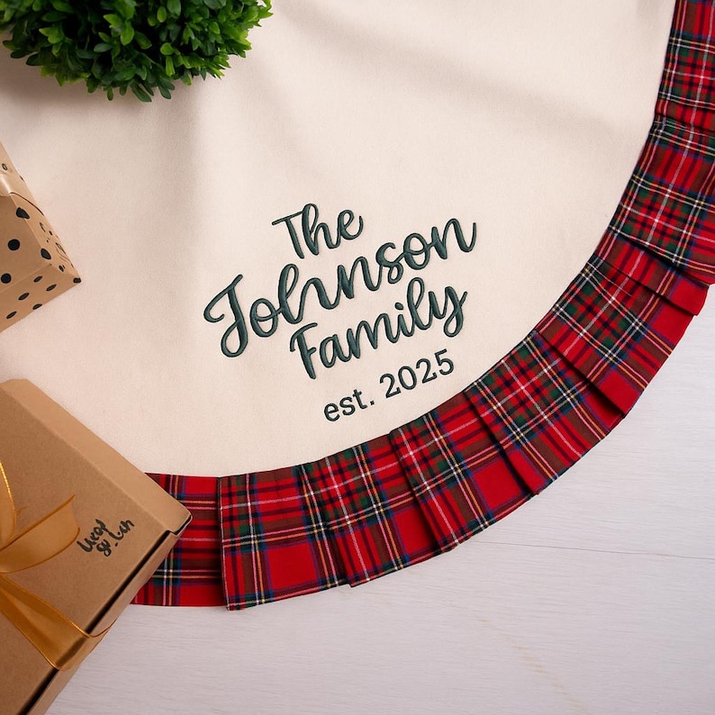 May include: Cream-colored Christmas tree skirt with a red and green plaid border. The skirt is embroidered with "The Johnson Family est. 2025" in green script. A gift box and greenery are visible in the background.
