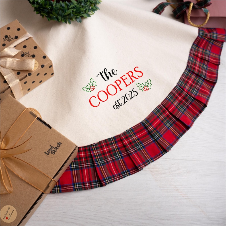 May include: Cream-colored Christmas tree skirt with a red and green plaid ruffle trim. The skirt features the text "The COOPERS est.2025" in black and red. Two wrapped gifts are visible in the image.
