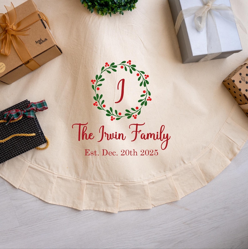 May include: A cream-colored Christmas tree skirt with a ruffled edge. The skirt features a wreath design with the letter "J" in the center, and the text "The Irwin Family Est. Dec. 20th 2025" in red. Several wrapped gifts are arranged around the skirt.