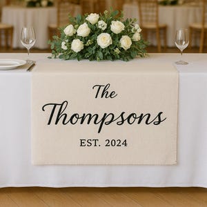 Puede incluir: Un camino de mesa color crema con el texto "The Thompsons EST. 2024" en escritura negra. El camino de mesa está colocado sobre un mantel blanco, con un arreglo floral y copas de vino en el fondo. El escenario parece ser una recepción de boda.